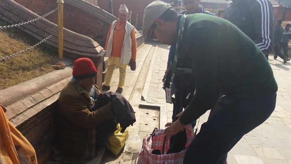 A volunteer crouches to pass a bag to a man in a red cap sitting at the edge of a paved walkway.