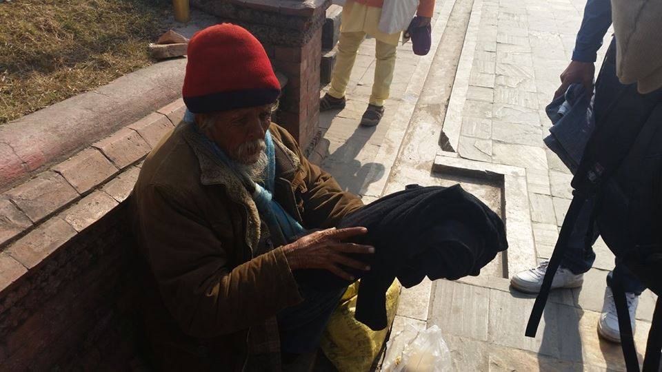 A man in a red cap sitting on a ledge receives a folded dark garment, passers by walking behind him.