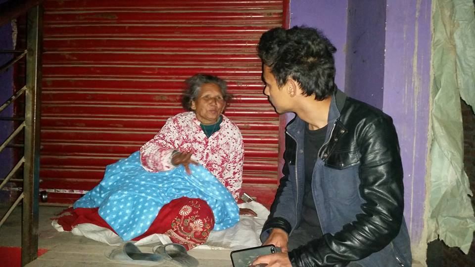 A volunteer crouches beside an elderly woman sitting in a shop doorway at night, a blanket spread over her legs.