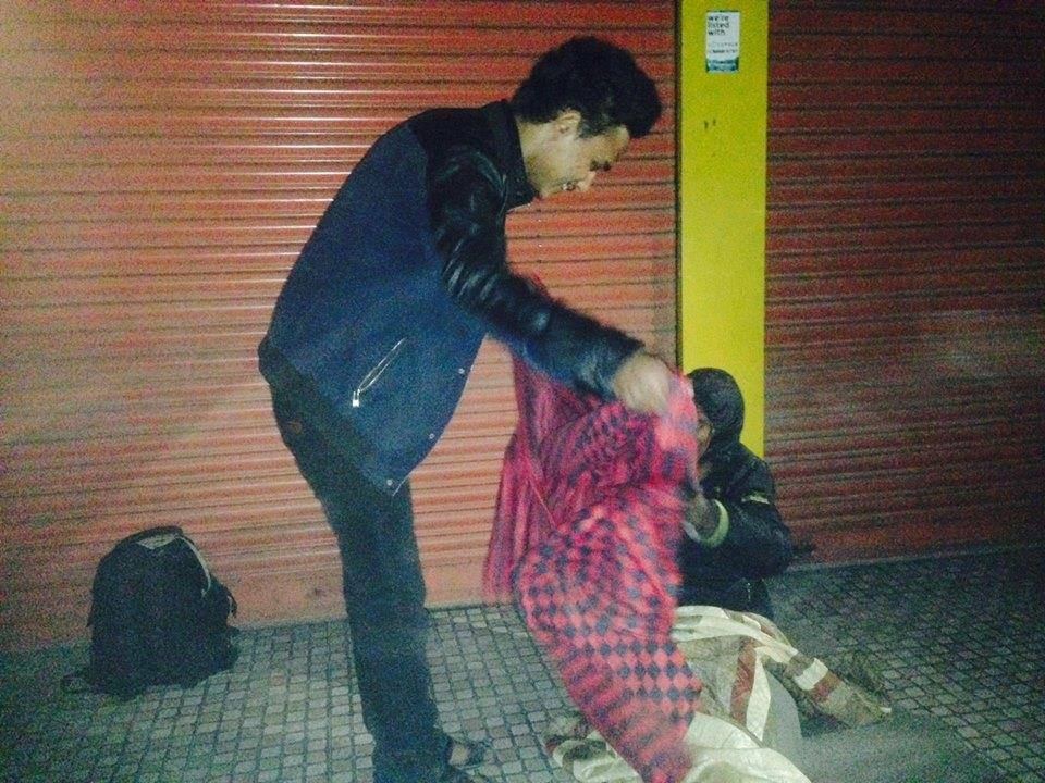 A volunteer bends to lay a blanket over a person lying against a closed shop shutter on a tiled pavement.