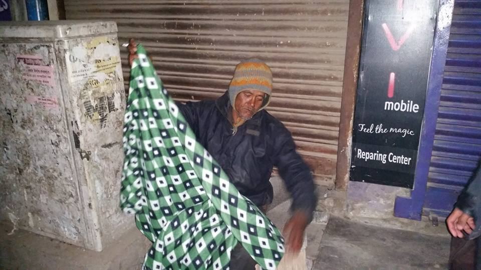 A man sitting on a pavement at night pulls a green patterned blanket around himself beside a closed shop.