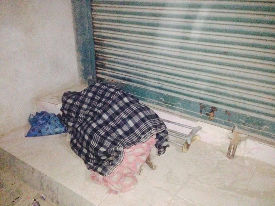 A person lies asleep on a low ledge in front of a closed shutter, covered by a checked blanket.