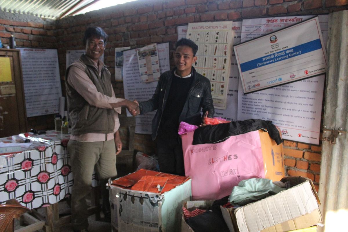Two men stand beside stacked cartons and piles of folded clothes inside a brick room, with printed charts pinned to the wall behind them.