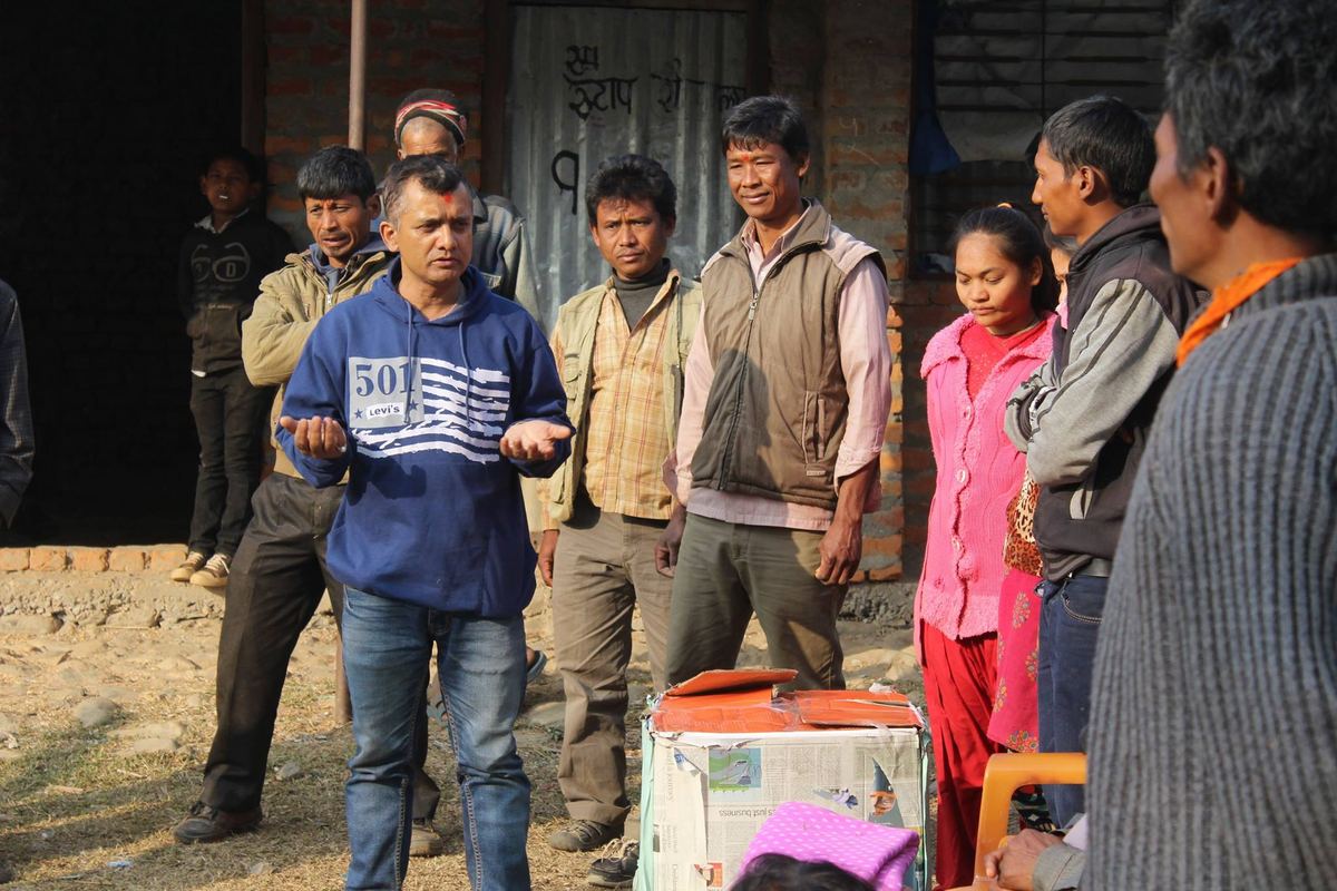 Villagers stand in a group outside a corrugated iron building around an open bundle of clothing on the ground.