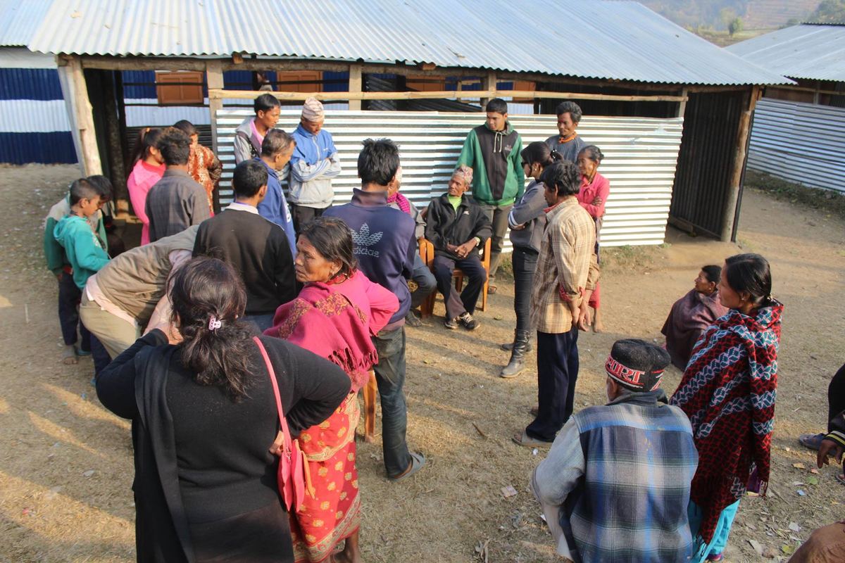 A crowd of adults and children stand and sit on a bare dirt yard in front of tin roofed shelters, waiting.